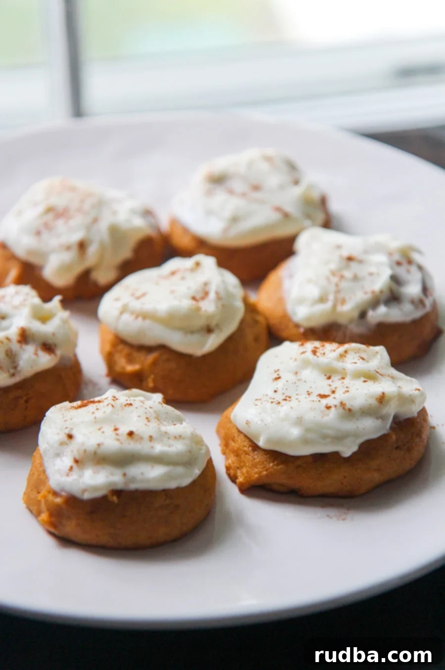 A close-up of a plate filled with perfectly baked pumpkin cookies, each topped with a generous swirl of cream cheese frosting and a light dusting of cinnamon.