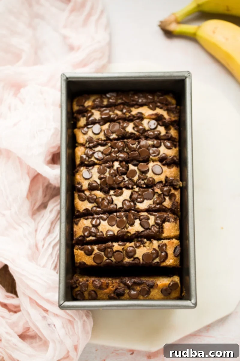 banana bread in a loaf pan cut into slices with bananas and a pink towel on the side