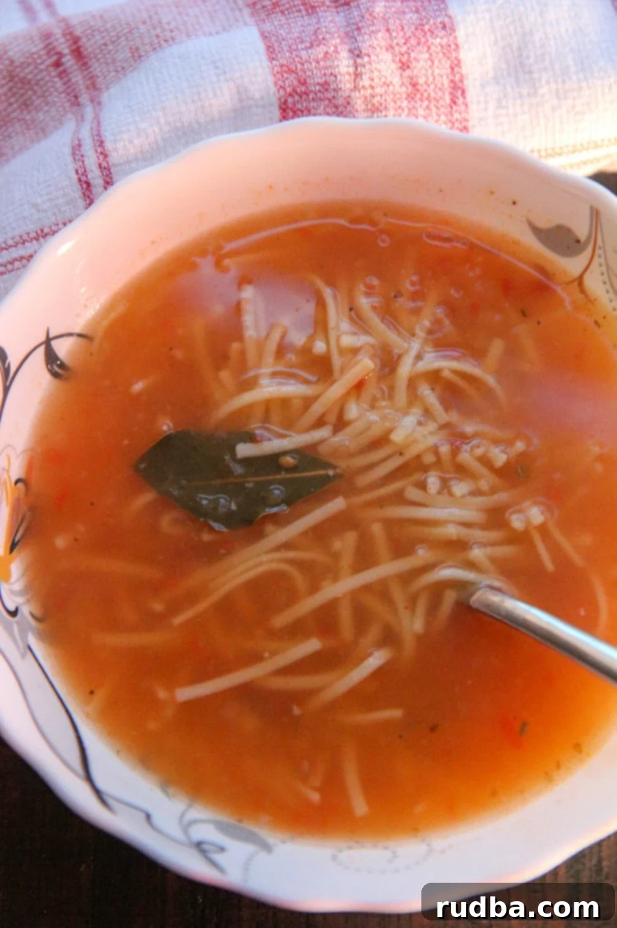 A vibrant bowl of Sopa de Fideo, garnished with fresh cilantro and a squeeze of lime, with a spoon resting in the rich broth and a red and white checkered towel on the side, ready to be enjoyed.