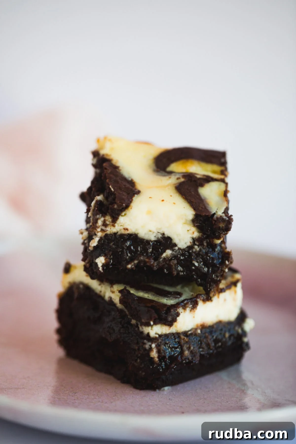 Two cheesecake brownies stacked on top of a pink plate, showing the rich texture.