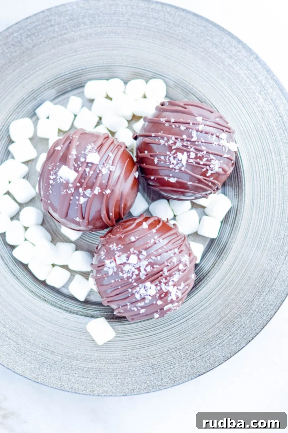 three hot chocolate bombs on a plate with mini marshmallows and a drizzle of caramel