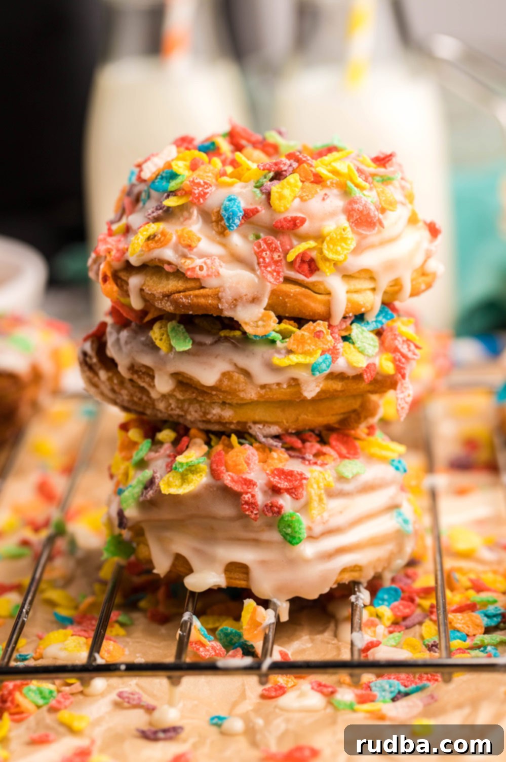 Three golden-brown air fryer fruity pebbles donuts stacked on a wire cooling rack, with a blurred glass of milk in the background, inviting a delicious breakfast or dessert experience.