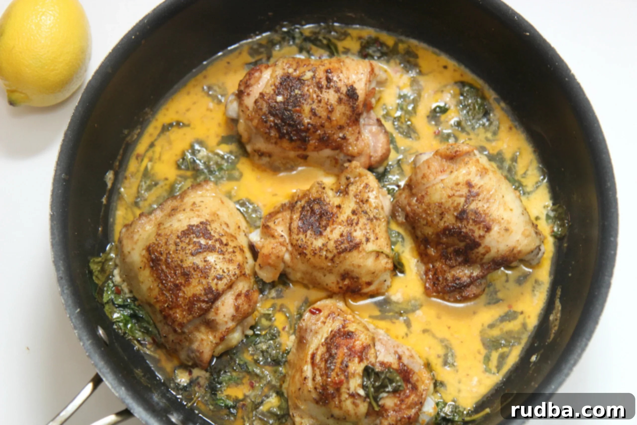 Five bone-in chicken thighs nestled in a rich, creamy yellow lemon spinach sauce in a black cast iron skillet, garnished with fresh herbs and a lemon half.