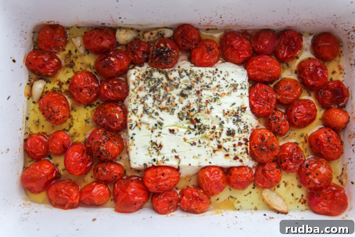 a block of feta cheese, olive oil, Italian seasoning, and cherry tomatoes arranged in a white baking dish before baking
