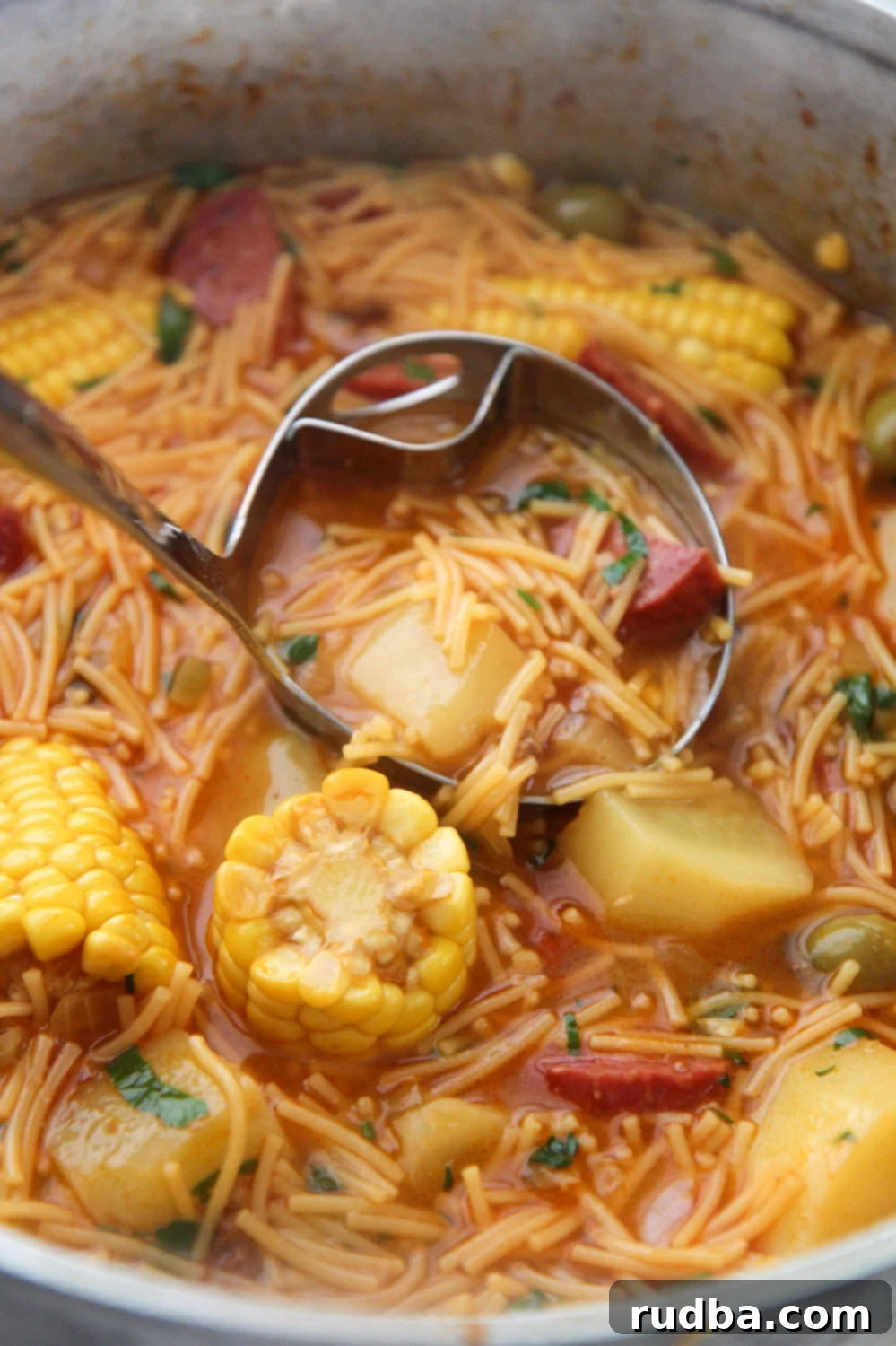 A ladle scooping hearty salami soup from a large pot, rich with vegetables and noodles.