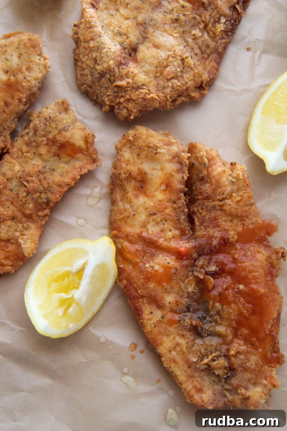 A close-up of a fried tilapia fillet with fresh lemon wedges and a small bowl of hot sauce on the side.