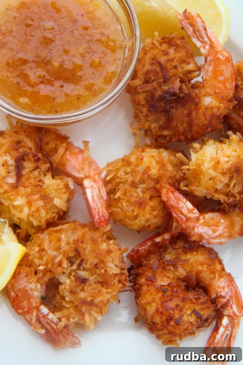 Six golden-brown fried coconut shrimp arranged on a plate with a side of orange dipping sauce and fresh lemon wedges.