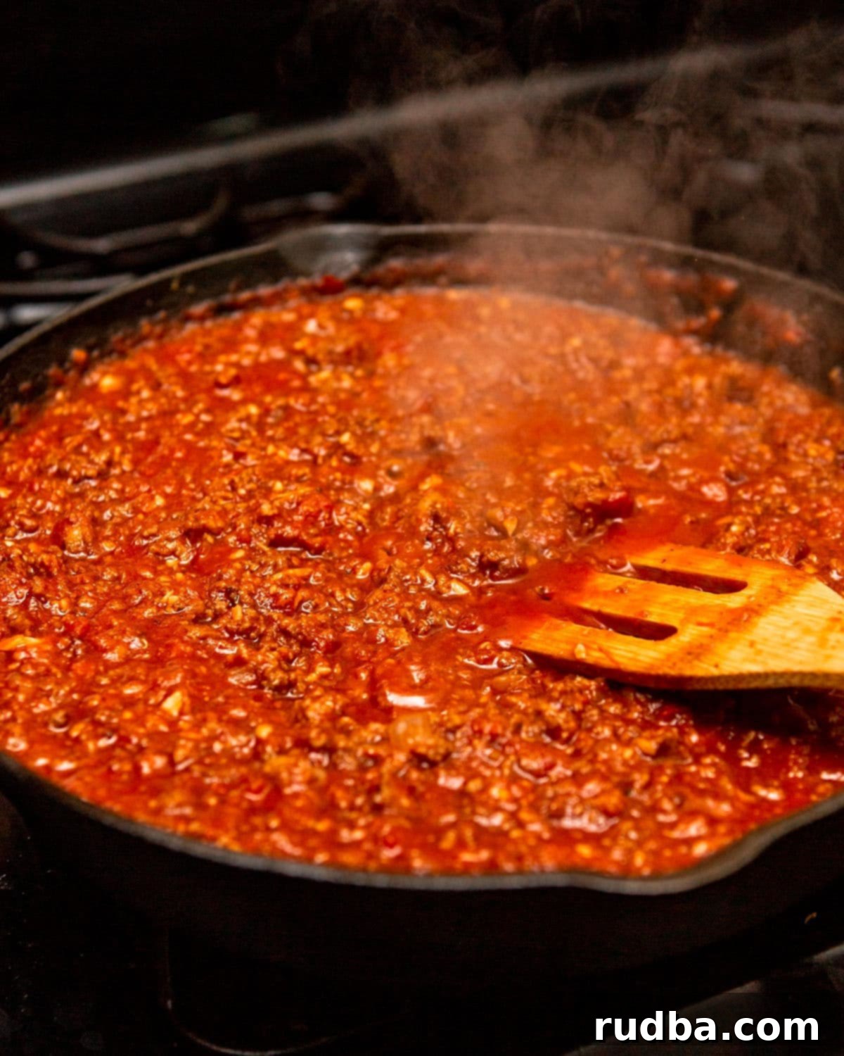 red meat sauce simmering in a cast iron skillet with a wooden spoon.