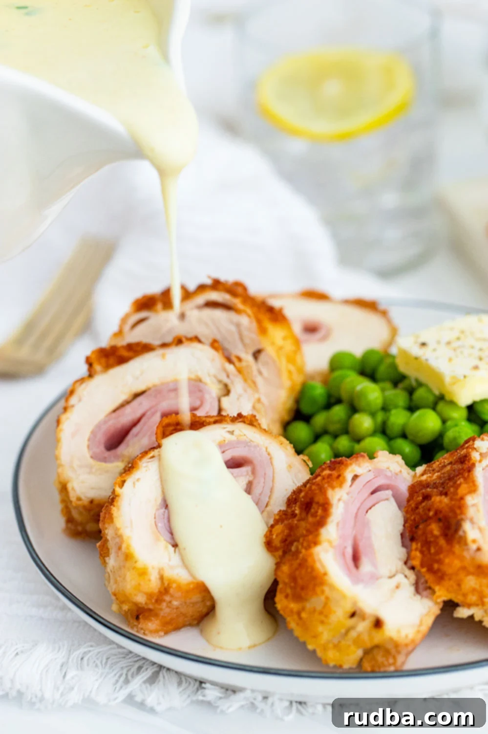 Perfectly cooked chicken cordon bleu on a white plate, drizzled with creamy Dijon sauce, accompanied by fresh green peas.