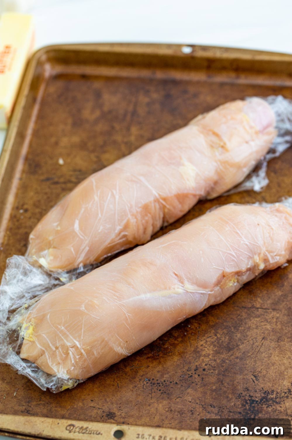 Two chicken breasts, tightly rolled with ham and cheese, secured in plastic wrap and placed on a baking sheet, ready for chilling.
