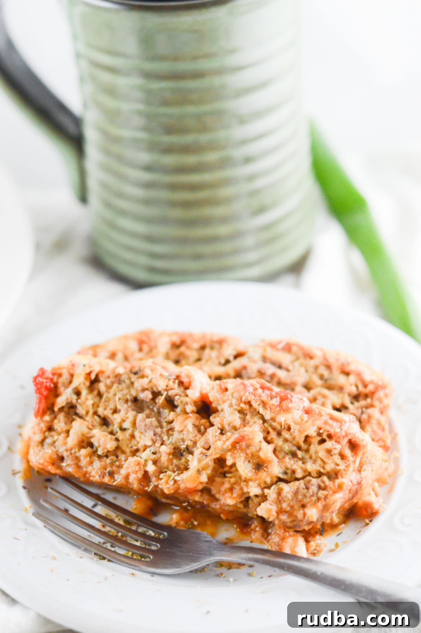 two slices of meatloaf on a white plate with a fork and a cup in the background. 