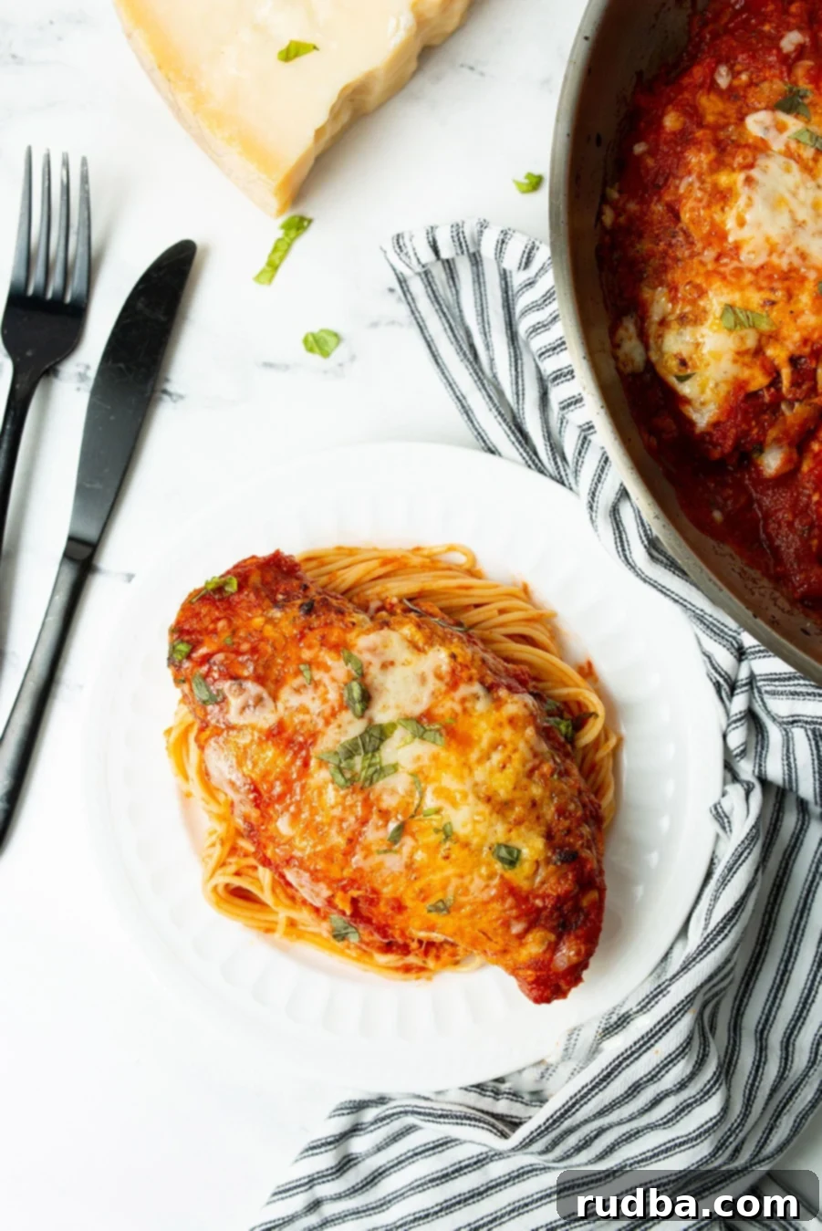 chicken parmigiana with spaghetti on a white plate with a knife and fork on the side and a blue and white towel. 