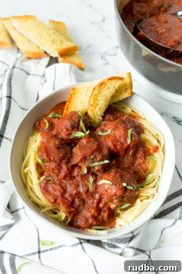 A white bowl filled with spaghetti and meatballs, garlic bread on the side, a white and blue towel underneath the bowl, and a pot of meatballs in the background.