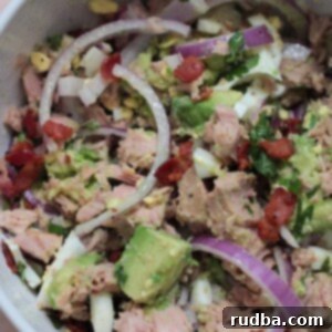 A vibrant bowl of creamy avocado tuna salad with visible pieces of avocado, tuna, bacon, and egg, garnished with fresh cilantro.