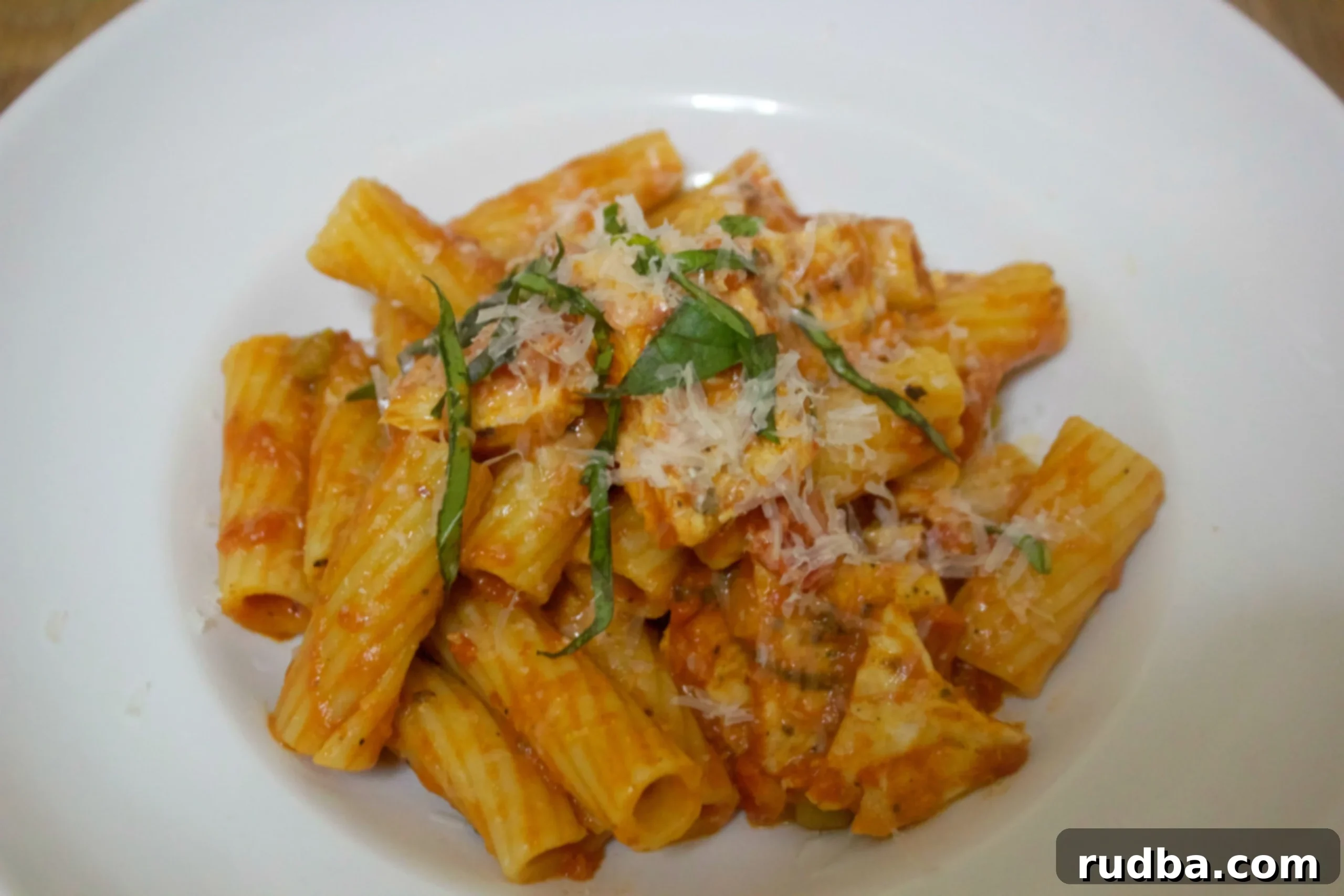 Spicy chicken, rigatoni, parmesan cheese, and fresh basil in a white bowl. 