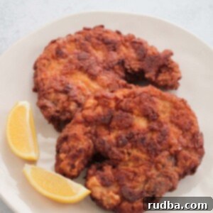 two fried pork chops on a plate with two lemon wedges.