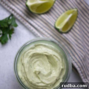 Closeup of creamy avocado crema in a small jar, garnished with a sprig of cilantro, with blurred limes and avocados in the background.