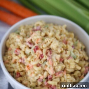 A large white bowl filled with creamy deli-style macaroni salad, garnished with fresh herbs. Carrots and celery are blurred in the background, suggesting fresh ingredients.