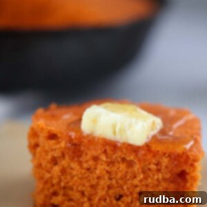 Golden Sweet Potato Cornbread 2 A square slice of golden sweet potato cornbread, topped with a dollop of melted butter and a drizzle of honey, served on a round plate.
