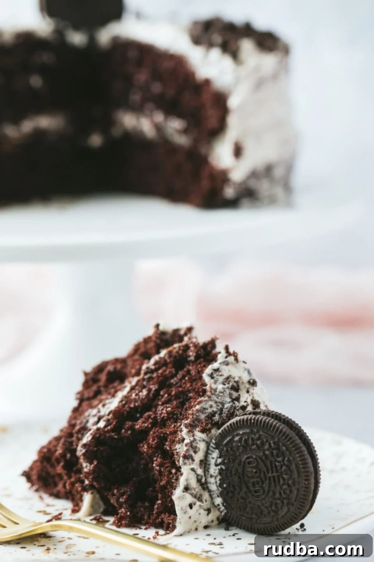 A perfectly sliced piece of Oreo cake on a white plate with a gold fork, with the remaining cake on a cake stand in the soft-focus background.