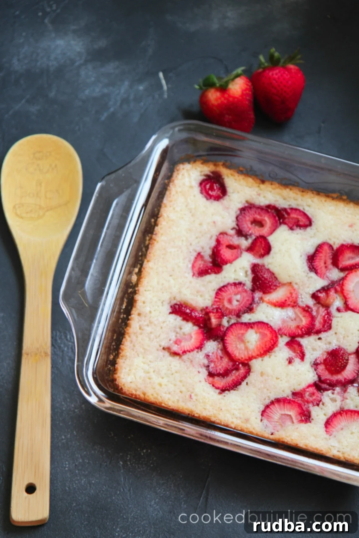 Fresh Strawberry Cobbler in a baking dish with a wooden spoon and two strawberries on the side. 