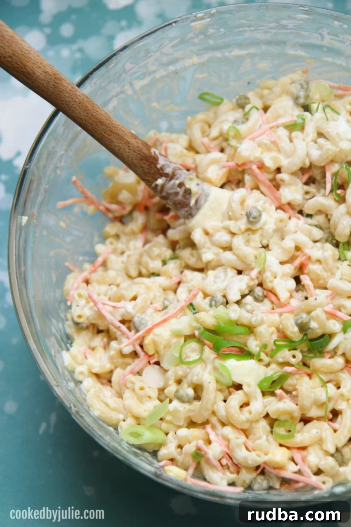 A large bowl of creamy Hawaiian macaroni salad, generously garnished with fresh green scallions on top. The salad showcases visible pieces of tender elbow macaroni, vibrant shredded carrots, and chopped hard-boiled egg, all coated in a rich, white dressing. The setting suggests a fresh and appetizing side dish.