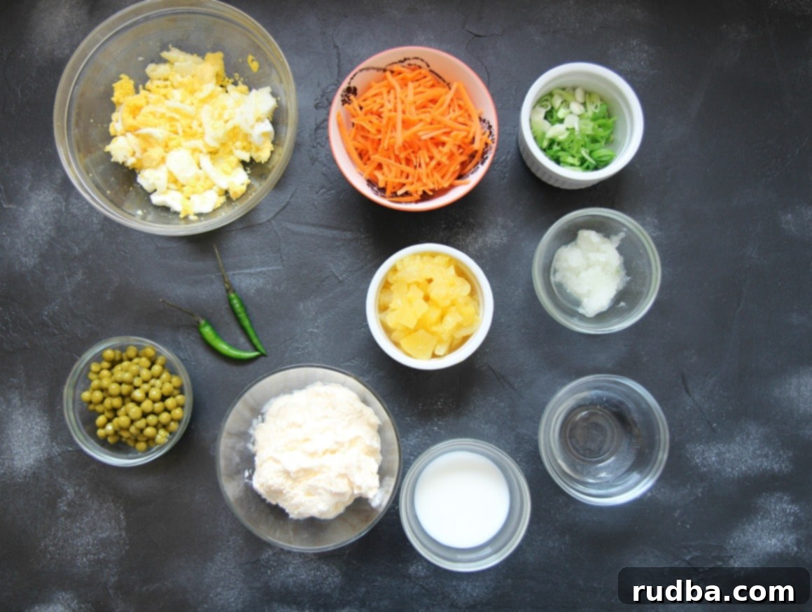 A flat lay photograph showcasing all the fresh and colorful ingredients ready for the Hawaiian Macaroni Salad. Arranged neatly are bowls of fluffy hard-boiled eggs, brightly shredded carrots, creamy mayonnaise, a small jug of milk, a bottle of vinegar, freshly chopped scallions, and golden pineapple chunks. Two vibrant green chilies are also visible, hinting at optional spice.