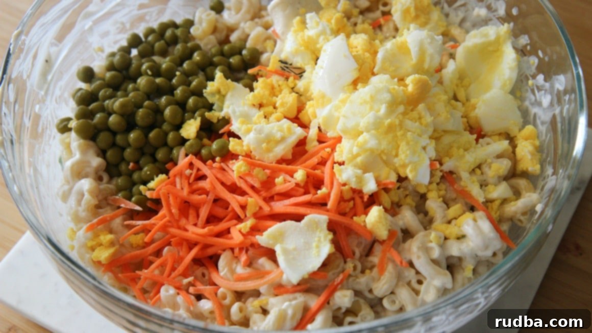 A vibrant close-up shot of a bowl filled with Hawaiian Macaroni Salad ingredients before mixing. Visible are tender elbow macaroni, bright green peas, shredded orange carrots, and chunky pieces of hard-boiled eggs, ready to be coated in the creamy dressing.