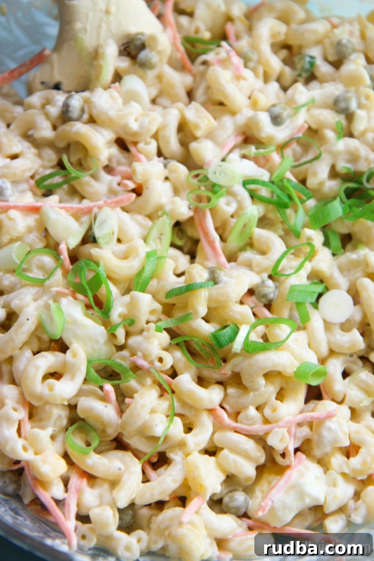 A close-up shot of a serving bowl filled with Hawaiian Macaroni Salad, artfully garnished with fresh green scallions on top. The image highlights the salad's luscious, creamy texture, with visible chunks of tender elbow macaroni, vibrant shredded carrots, and a hint of pineapple, all beautifully coated in the rich dressing.