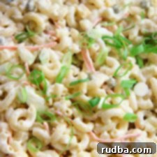 A vibrant close-up of a serving of Hawaiian Macaroni Salad, showcasing its creamy texture with visible elbow macaroni, shredded carrots, and a sprinkle of fresh green scallions on top. The salad is presented in a simple white bowl, ready to be enjoyed as a refreshing side dish.