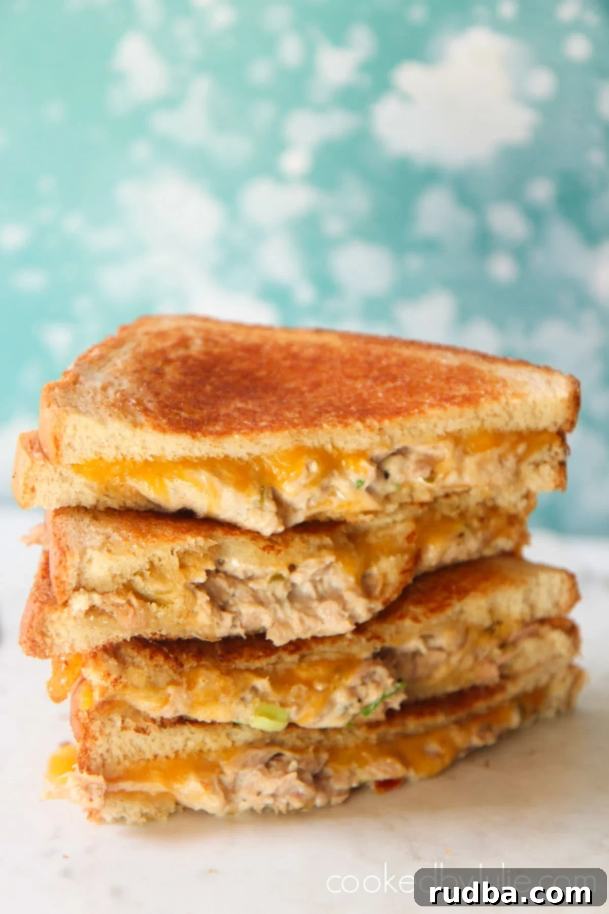Four golden-brown tuna melt sandwiches with melted cheddar cheese on a white surface, set against a baby blue and white backdrop. They look perfectly toasted and ready to eat.