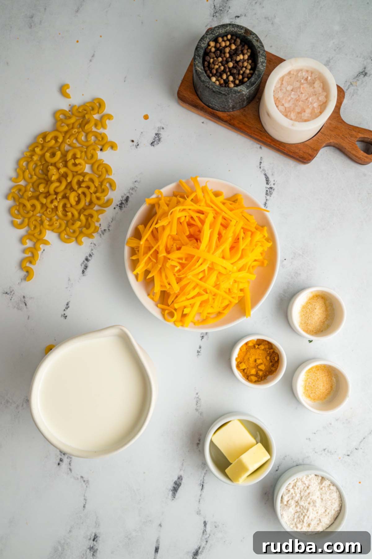 Arrangement of ingredients for homemade mac and cheese: shredded cheese, elbow macaroni, half and half, unsalted butter, and a selection of spices and flour on a clean white surface.