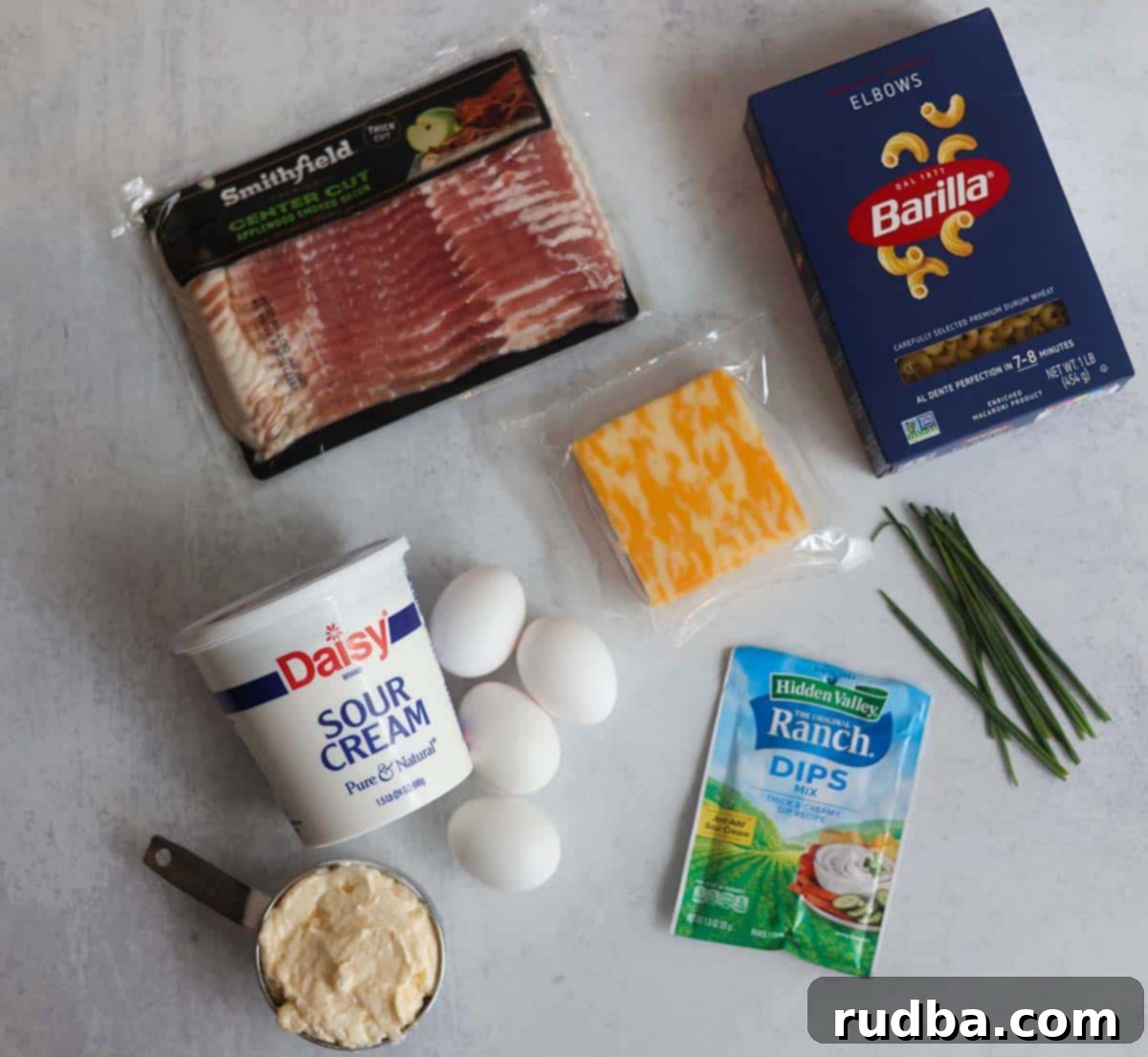 Assortment of fresh ingredients for creamy bacon ranch pasta salad, including uncooked elbow macaroni, a block of Colby Jack cheese, strips of raw bacon, a bunch of chives, and hard-boiled eggs, all neatly arranged.