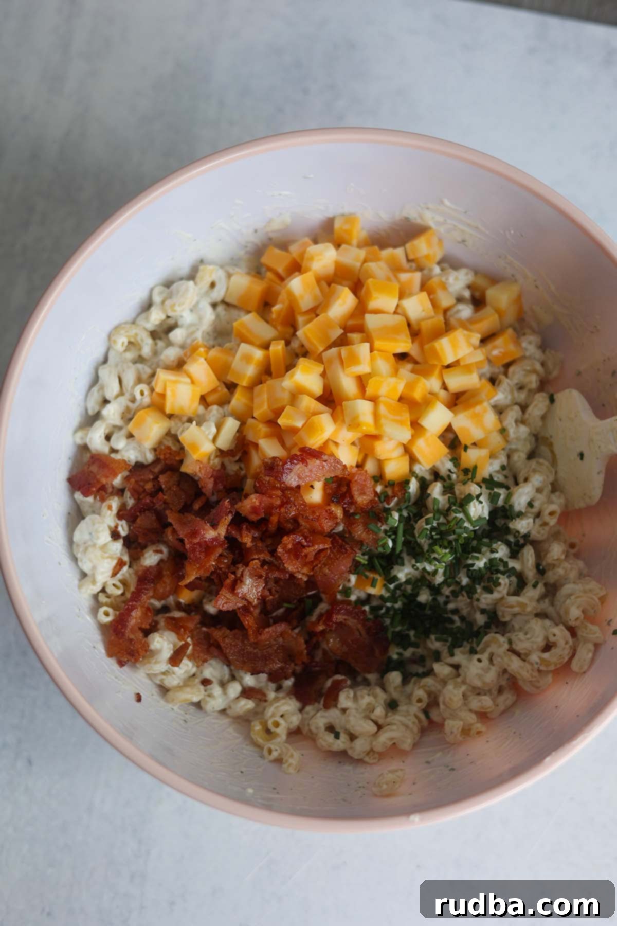 A large bowl containing cooled macaroni, diced Colby Jack cheese, chopped crispy bacon, and fresh chives, partially mixed with a spatula, waiting for the creamy dressing.