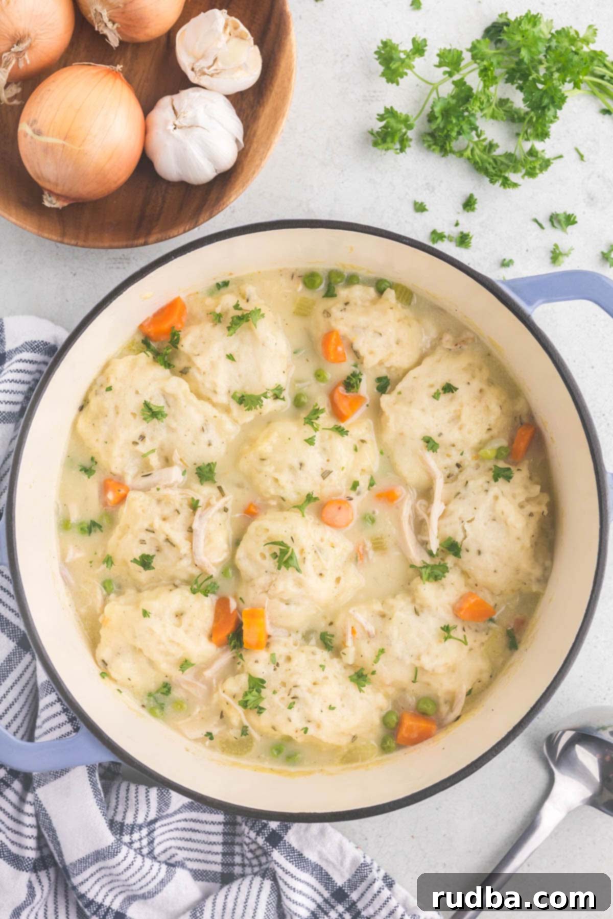 Hearty Homestyle Chicken and Dumplings 2 A pot filled with chicken and dumplings with onions, parsley, and garlic on the side.