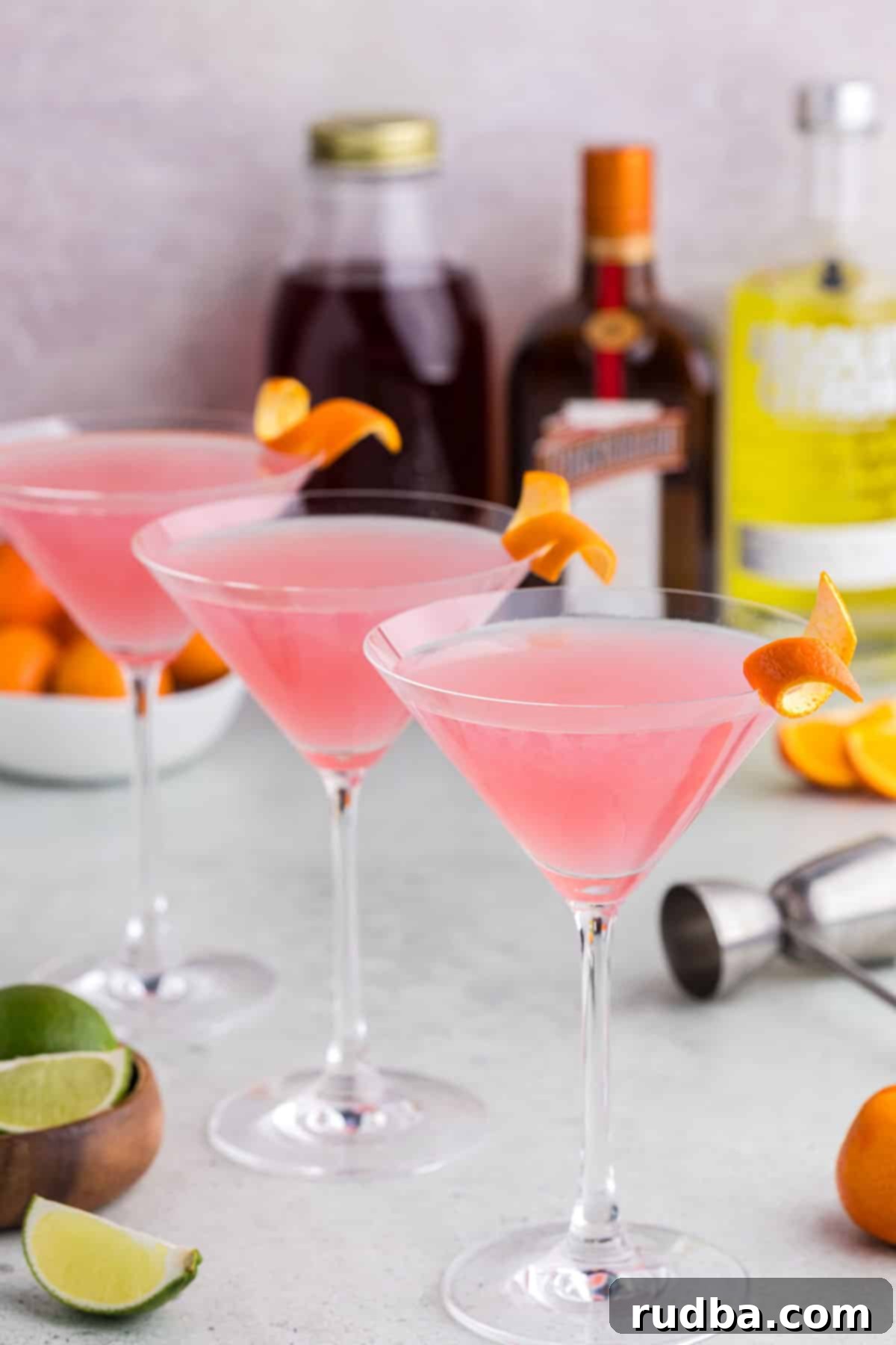 Classic Cosmopolitan Cocktail Recipe 2 Three cosmopolitan martinis with alcohol bottles in the background.