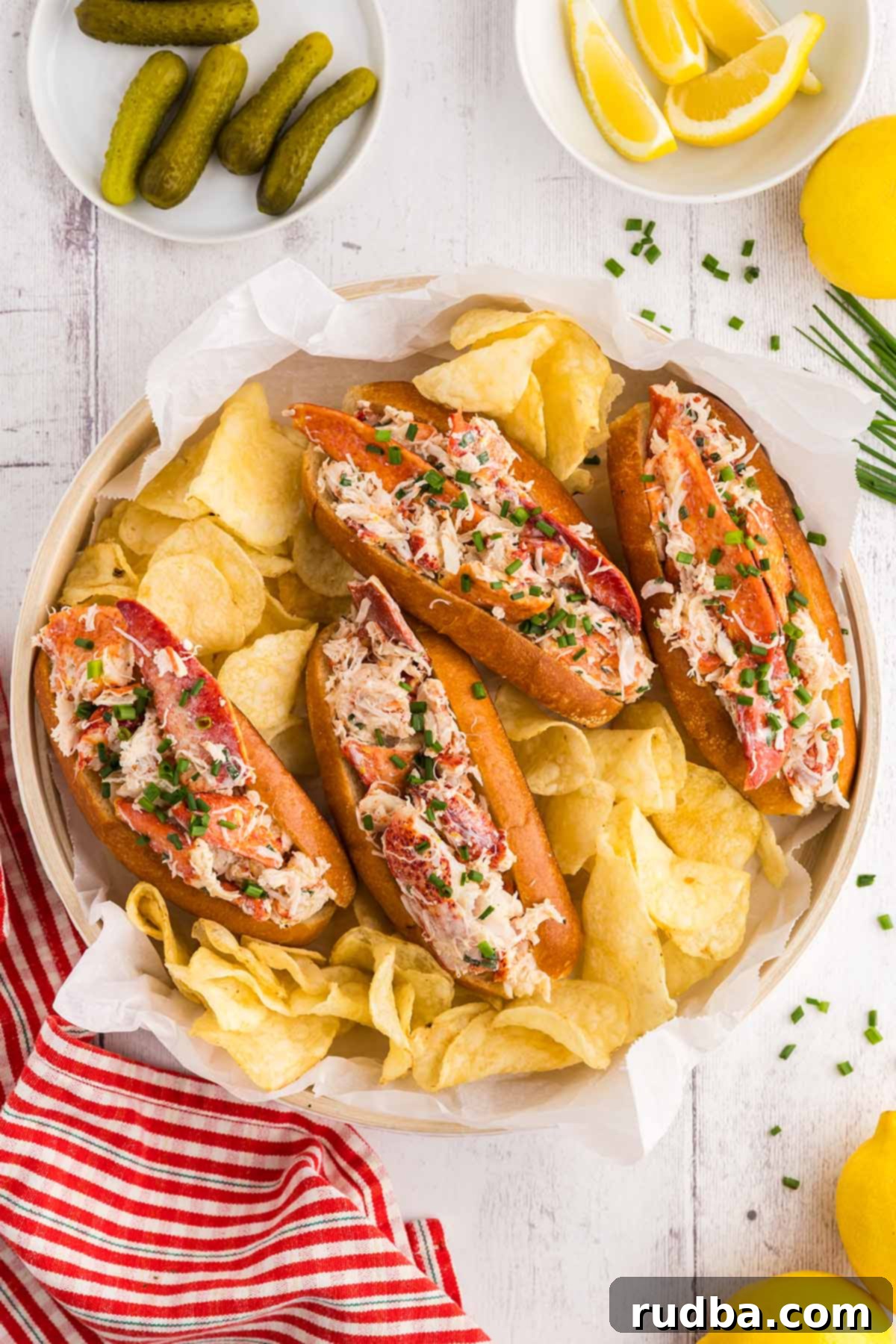 Four classic lobster rolls beautifully arranged with a side of golden potato chips, crunchy pickles, and vibrant lemon wedges, ready to be enjoyed.