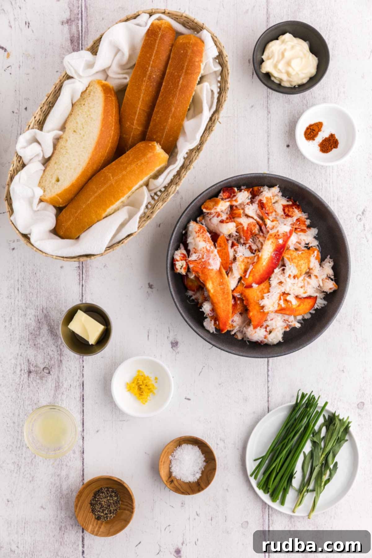 A selection of fresh ingredients laid out for making lobster rolls, including cooked lobster meat, mayonnaise, lemon, chives, tarragon, spices, and split-top buns.
