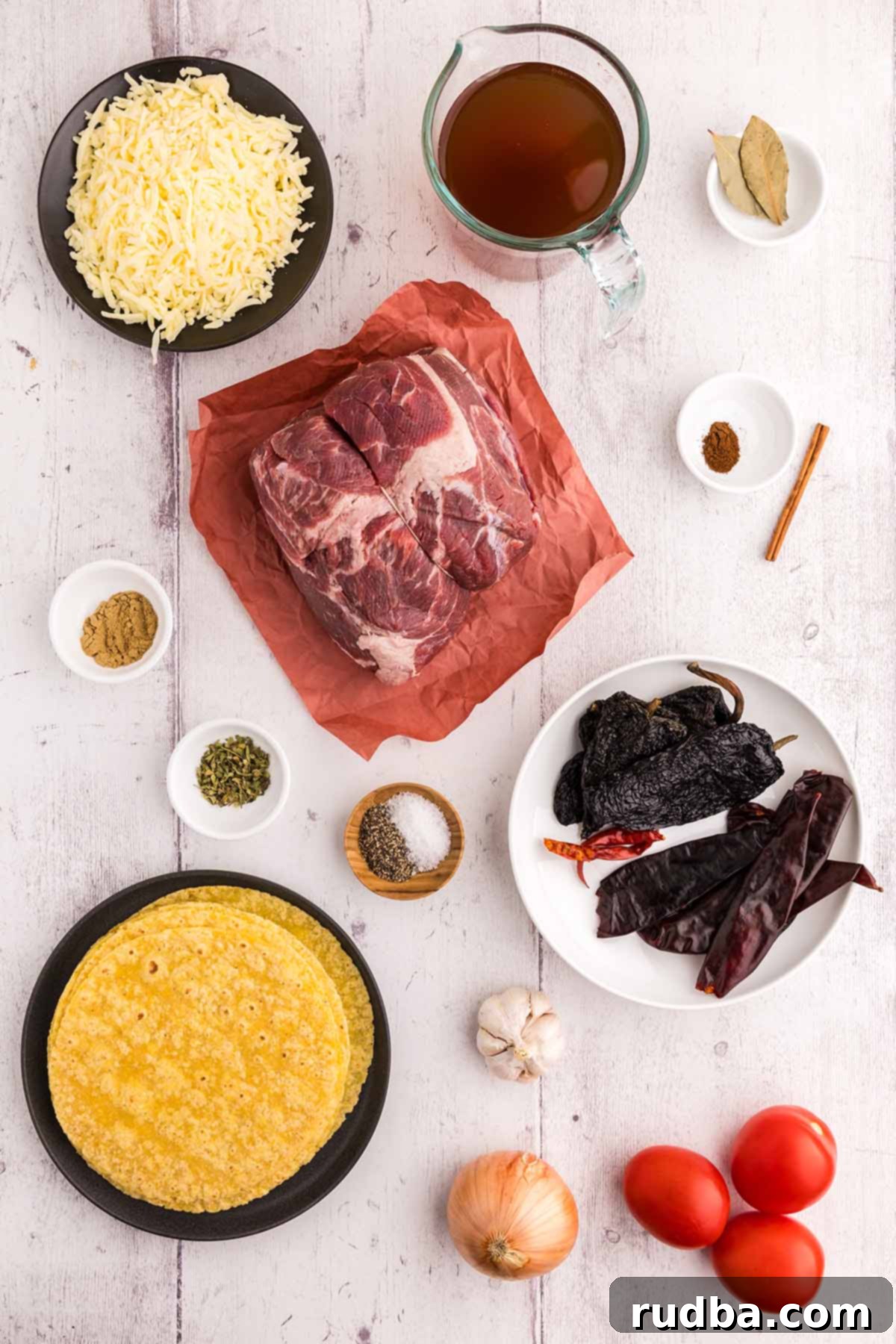 A vibrant flat lay showcasing all the essential ingredients for Birria Tacos: shredded cheese, beef broth, a variety of spices, bay leaves, raw chuck roast, corn tortillas, fresh tomatoes, onions, garlic, and a cinnamon stick arranged on a clean white surface.