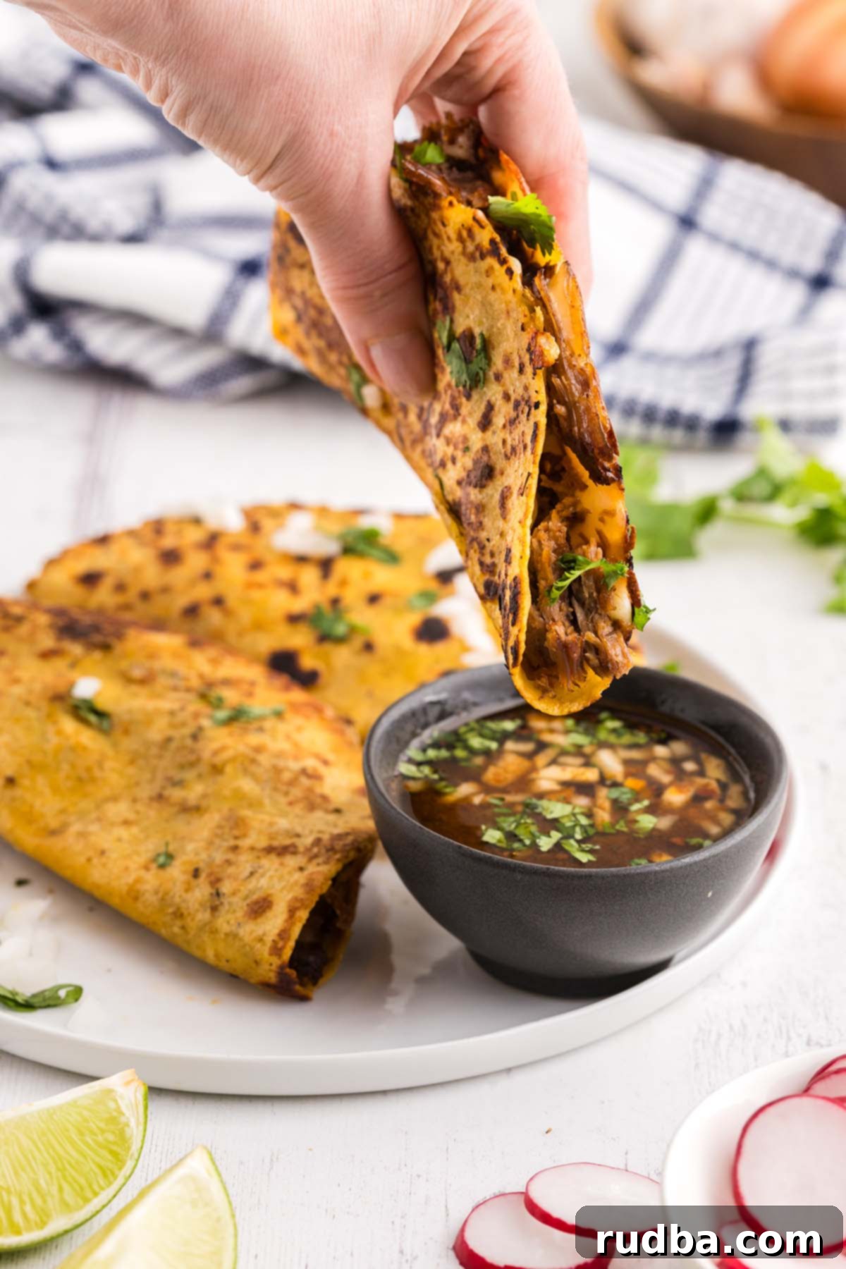 A close-up of a golden, crispy birria taco being dipped into a small bowl of rich, red consomé, illustrating the traditional way to enjoy this flavorful Mexican dish.