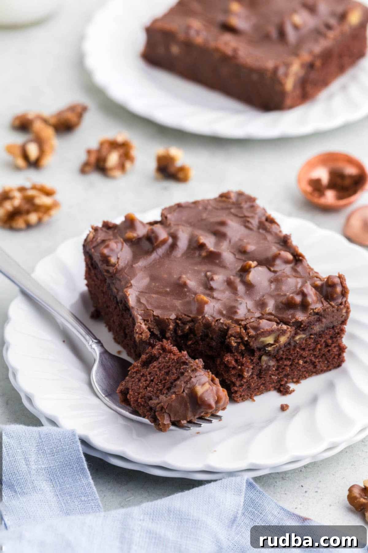 A perfectly portioned slice of Texas Chocolate Sheet Cake, rich with frosting and walnuts, resting on a pristine white plate beside a fork.