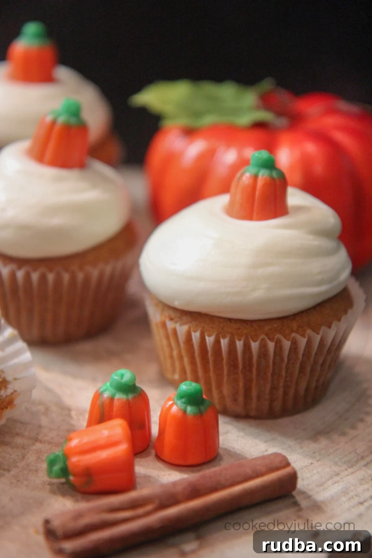 three pumpkin cupcakes with cream cheese frosting with a cinnamon stick, three pumpkin candies, and a fake pumpkin on the side. 