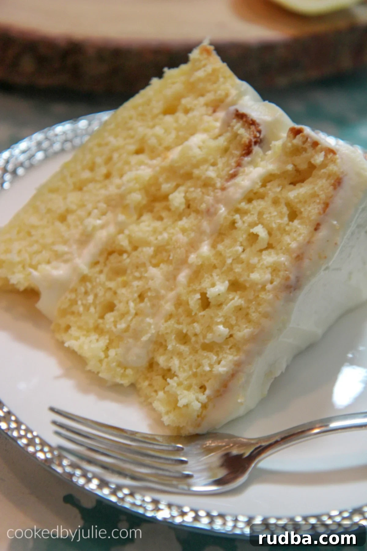 A beautifully sliced three-layer lemon cake on a white and silver plate with a fork on the side, showing its moist layers and creamy frosting.