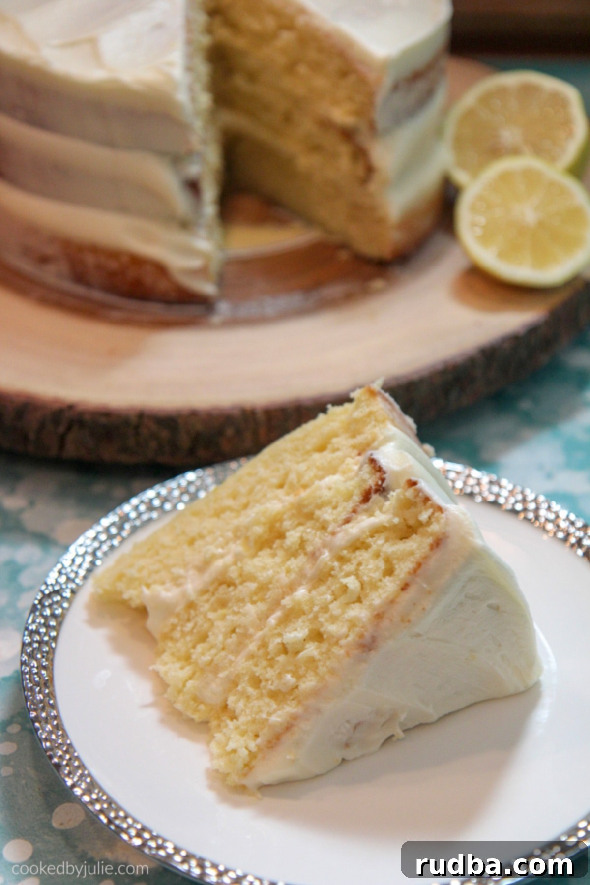 A vibrant lemon cake slice on a white and silver plate, with a full three-layer lemon cake in the background adorned with fresh lemon slices on a rustic wooden cake stand.