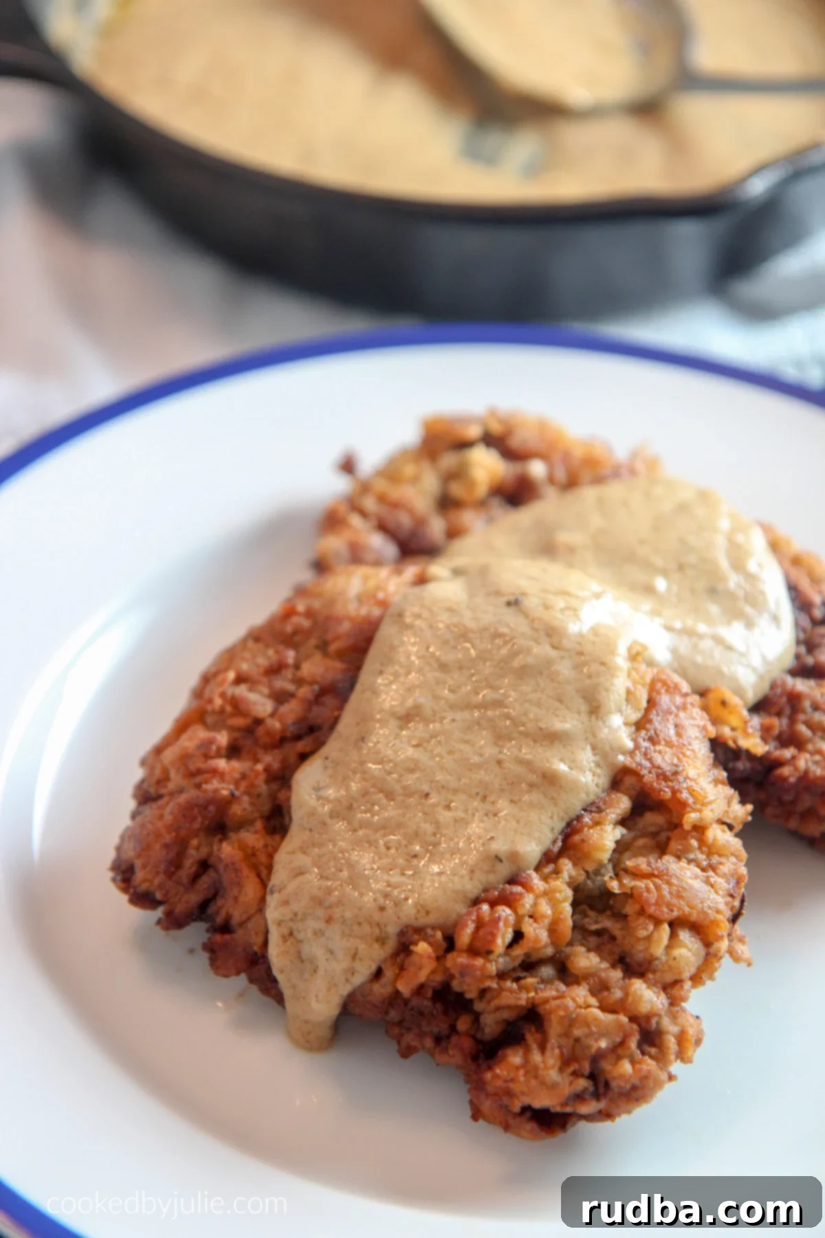 Two pieces of golden-brown chicken fried steak on a white and blue plate, generously covered with creamy white gravy. An iron skillet with more gravy and a serving spoon rests in the blurred background, hinting at the deliciousness to come.