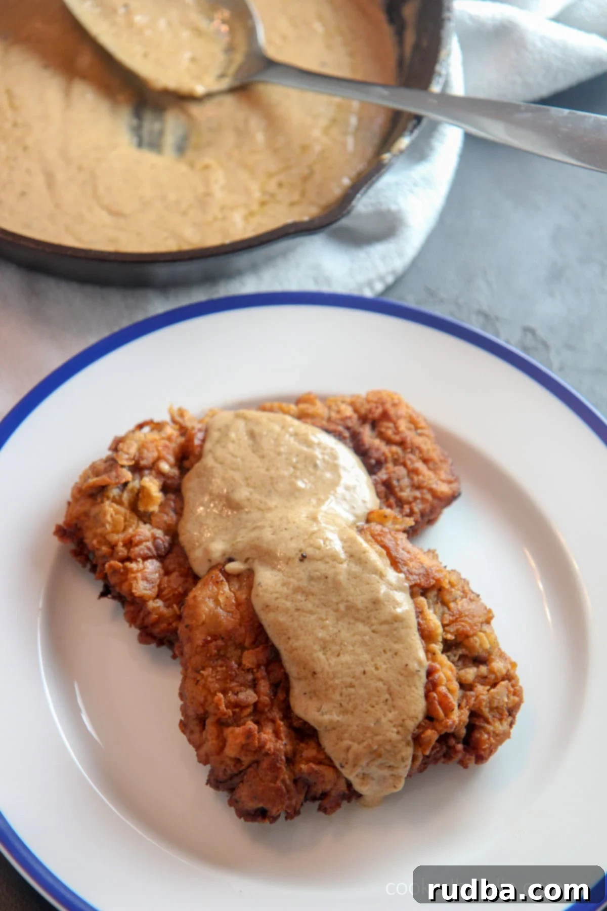 Two chicken fried steak cutlets perfectly plated with creamy white gravy on top, ready to be enjoyed. In the background, an iron skillet holds more gravy with a spoon, suggesting an abundance of this delicious sauce.