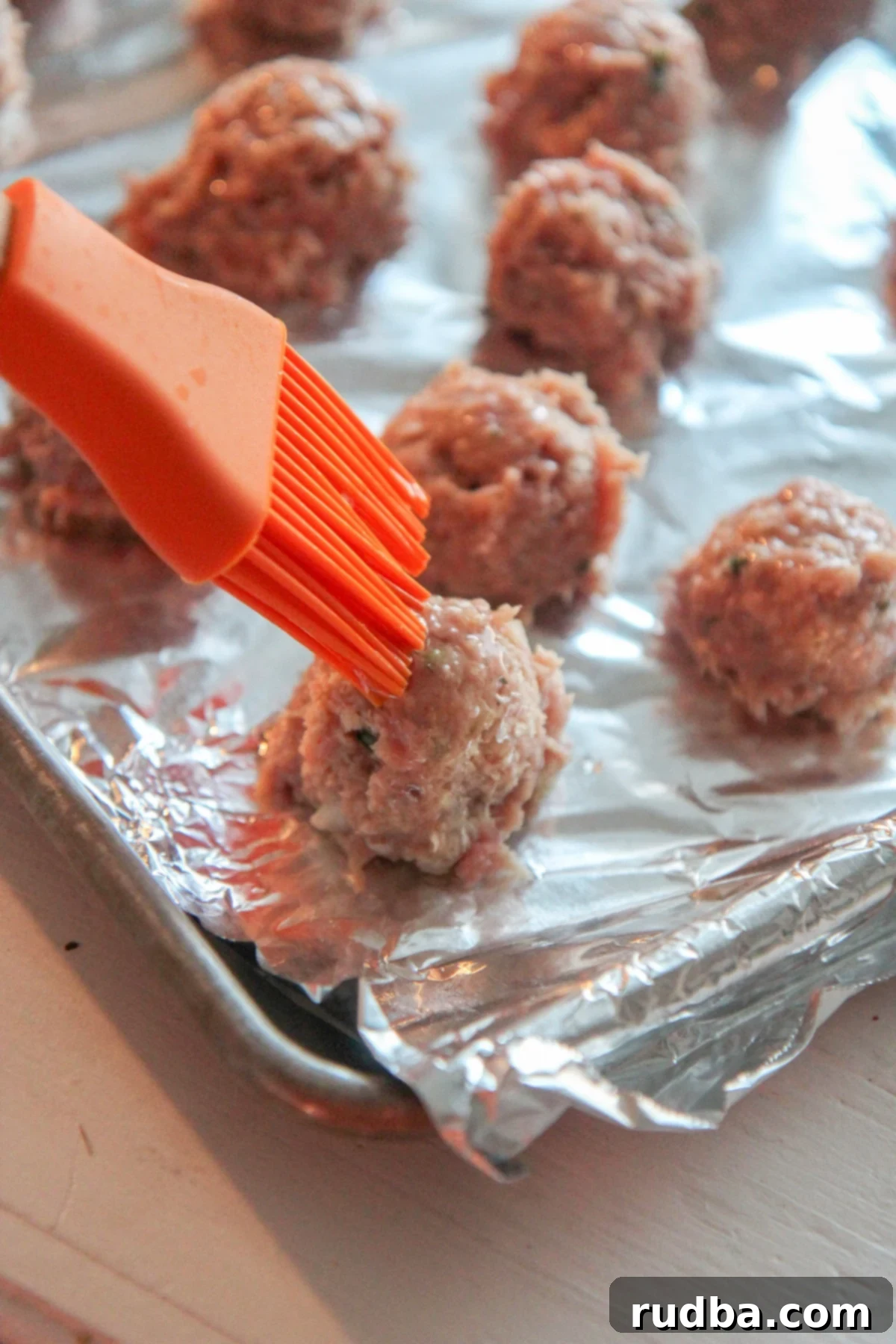 An orange pastry brush applying olive oil to uncooked turkey meatballs on a baking sheet.