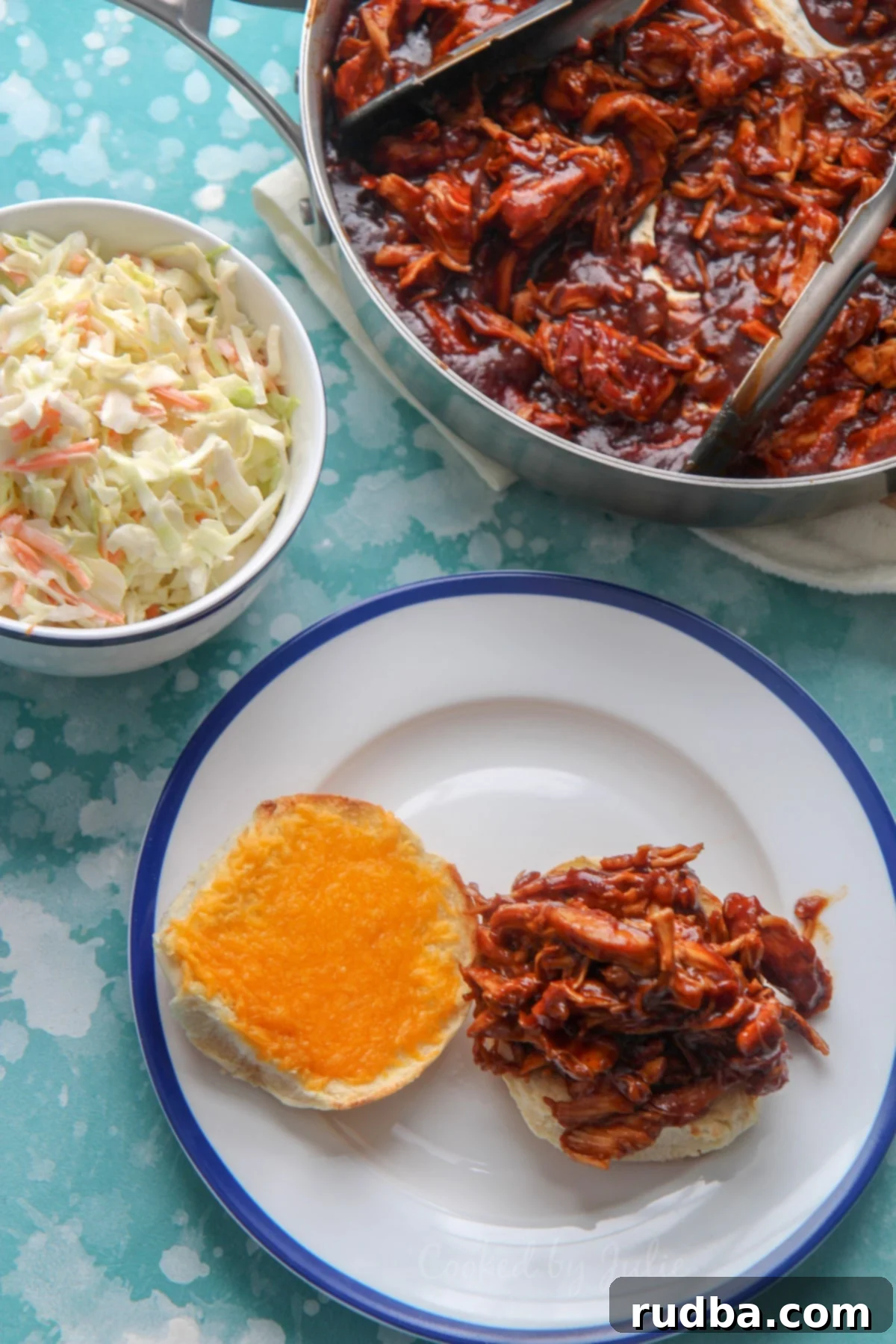 pulled chicken sandwich with cheddar cheese on a white and blue plate. Coleslaw in a bowl and pulled chicken in a skillet