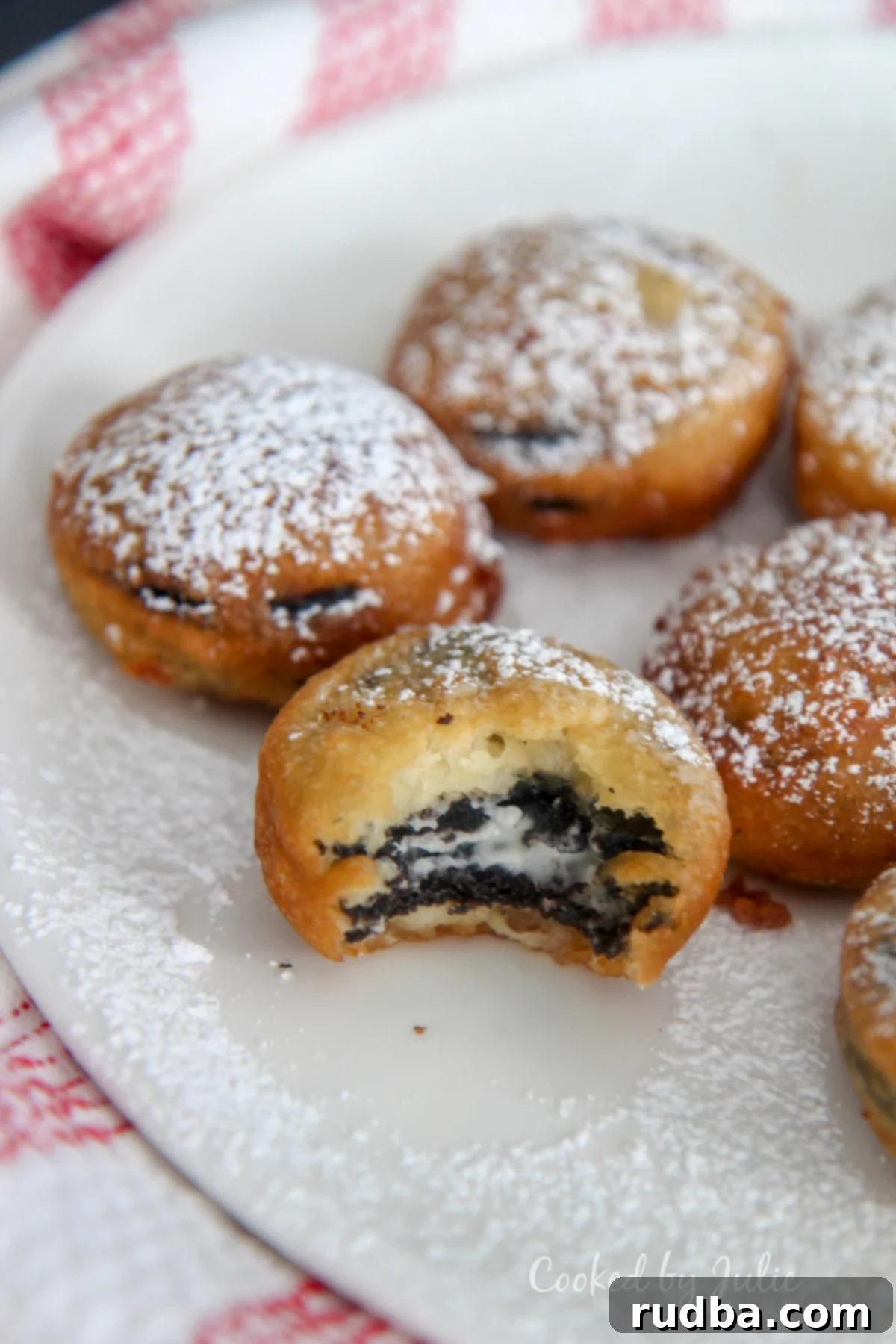 deep fried Oreos with powdered sugar on a white plate with one half eaten cookie and a red and white towel on the side. 