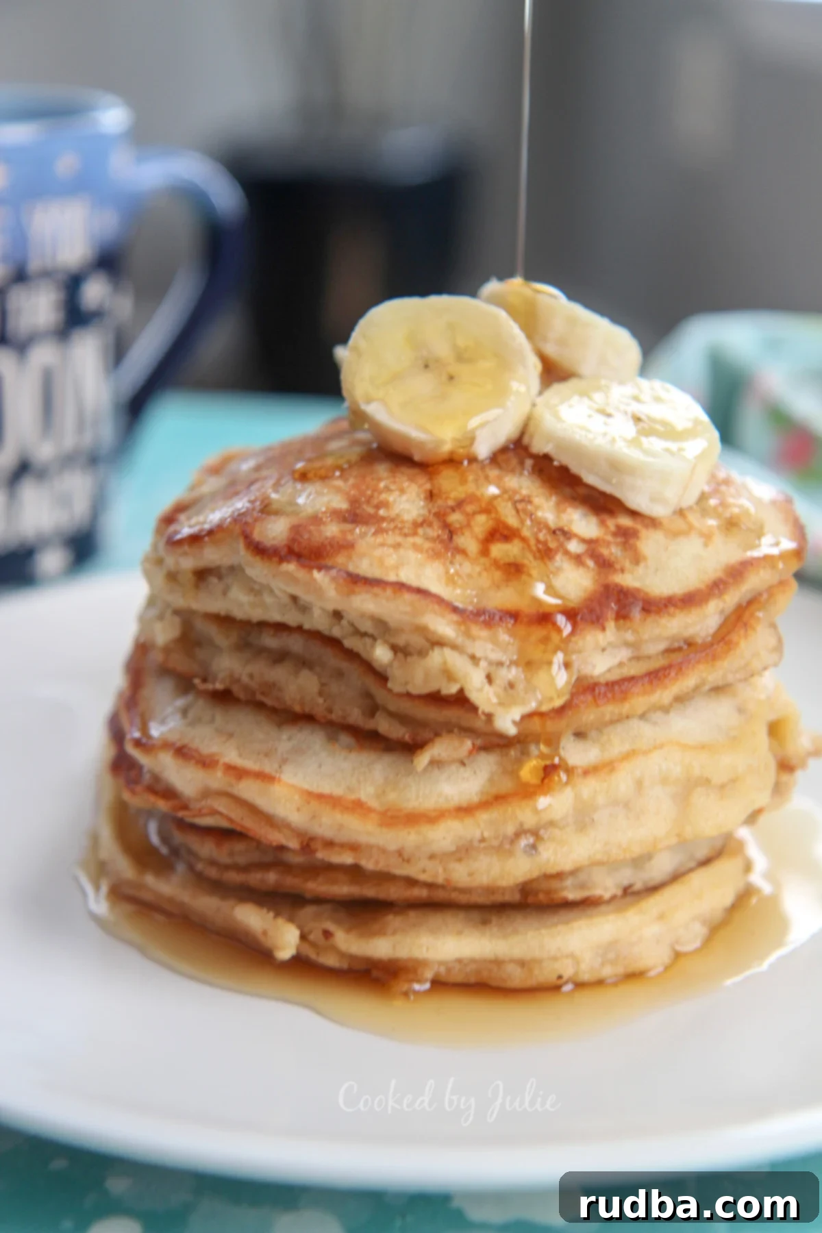 Six fluffy banana pancakes on a white plate with fresh banana slices on top, a generous drizzle of maple syrup, and a blue mug in the background.